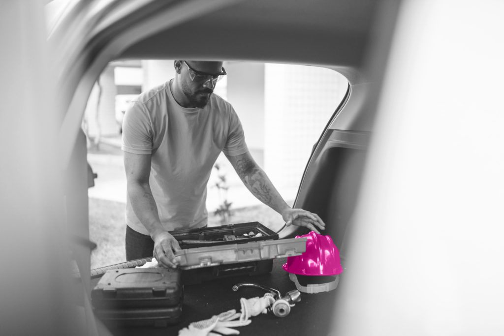 Black and white image of man putting a toolbox into the back of a tansit van. A helmet is placed next to the toolbox and is highlighted a magenta pink.