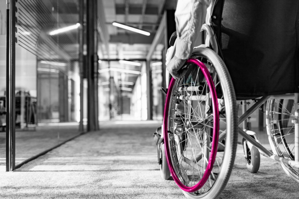A black and white image featuring a wheelchair user in the right-hand corner of the image in an office environment. The corridor is visible but no other people are in the building. The weelchair handle is coloured in a magenta pink which are the brand colours for Fraser Dawbarn with the user's hand looking to move forward through the corridor.
