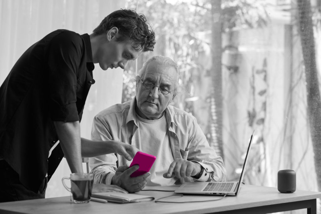 An black and white image of an old man sitting at a desk with a laptop in front of a glass window. He is showing a younger man his phone (which is coloured in pink) with a confused facial expression.