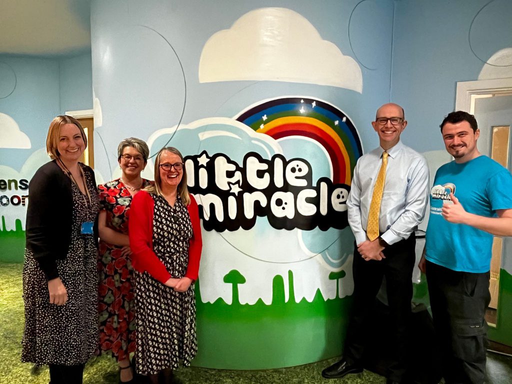 Image shows three Partners from Fraser Dawbarns and two staff members of Little Miracles at the Charity's head office in Peterborough. They are inside the building standing and smiling for a photo in front of a logo which has been printed onto a blue wall in the corridor of their building. From Left to right: Louise Evans, Helen Jarvis, Sarah Lamb, Iain Grimes, and Jack Cook.