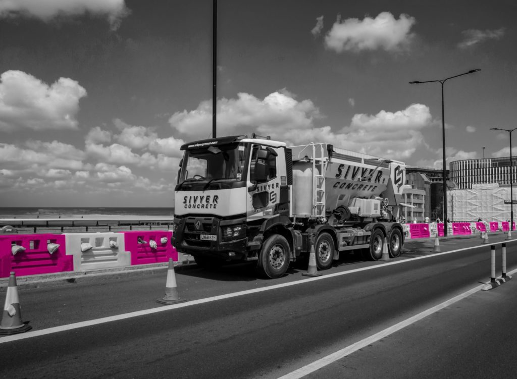 A black and white image of a construction lorry on a bridge in a city. There are warning cones around the vehicle as well as a temporary work barrier which has been coloured in pink and white to match the brand's colour palette.
