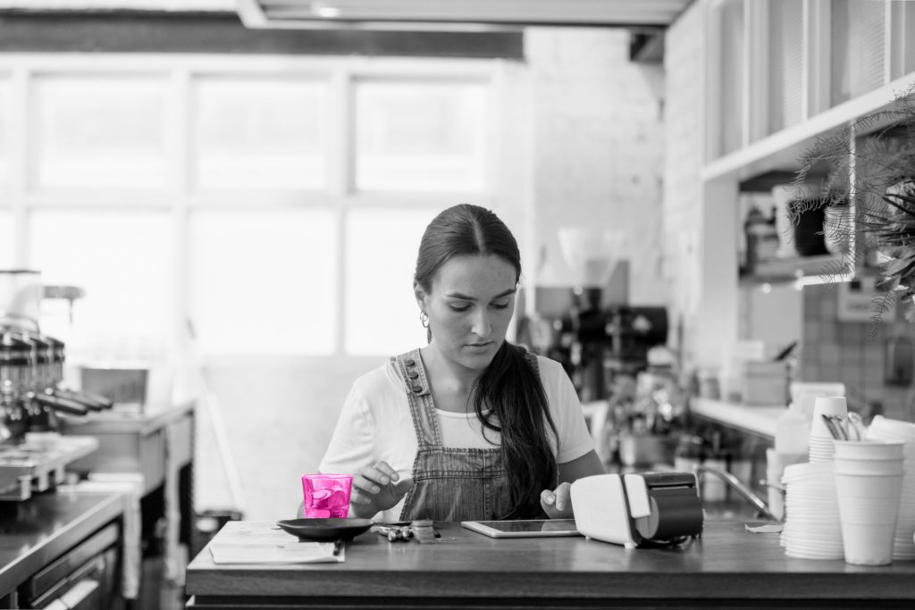 Black and white image of a young woman in a shop sat at a table looking at a piece of paper with a phone, card machine, and tip jar full of coins on the table.
