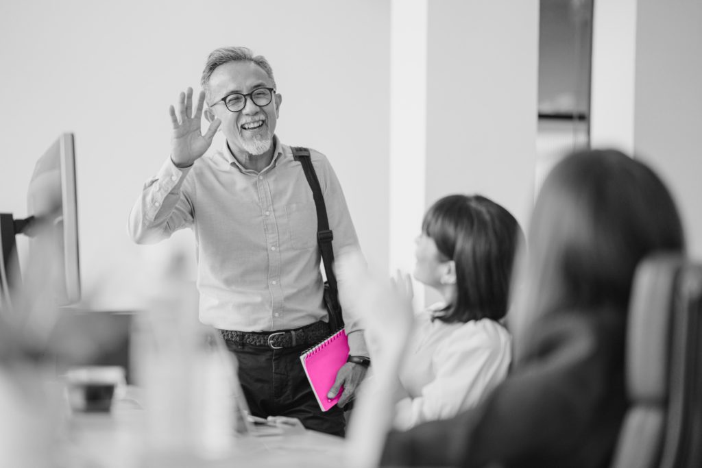 Image shows black and white office space with elderly man in glasses waving goodbye to younger colleagues. He is holding a bright pink notepad and carries a shoulder bag.