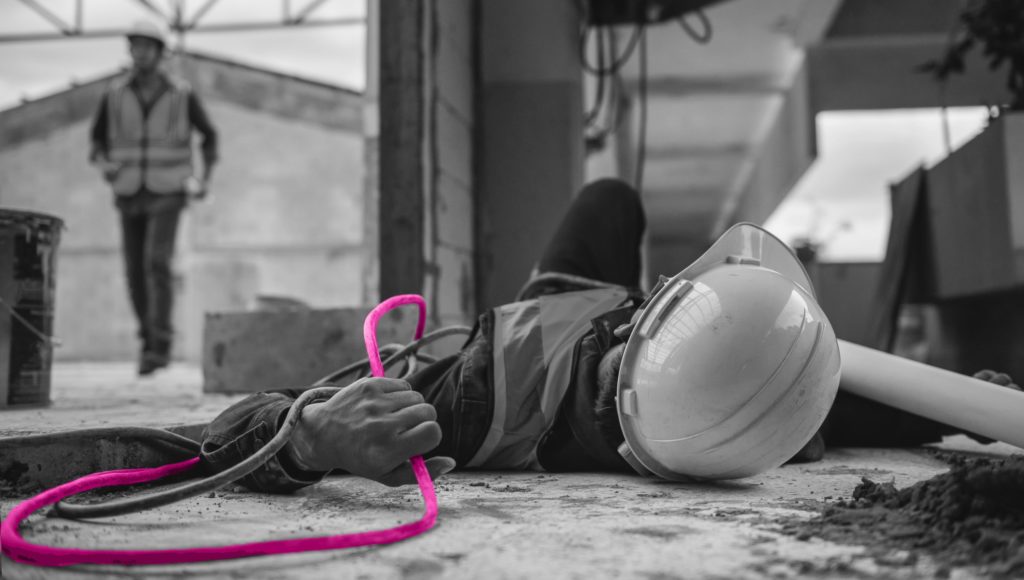 A black and white image showing a man in a high visibility jackets and hard hat who has fallen over in a construction setting holding a pipe. The pipe is coloured in a bright pink and there is another workman in the corner of the image overseeing the incident.