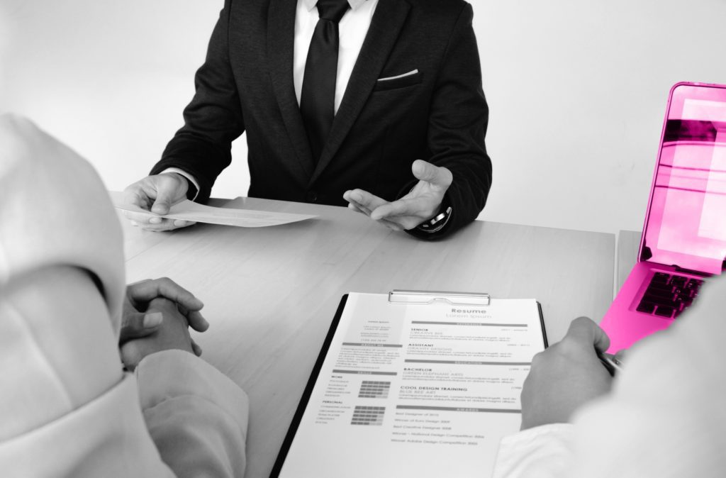 Black and white image of a man in a suit sat in front of two recruiters at a table. There is a laptop in front of the employer on the right (male) and an application form between them and the other employer (demale).
