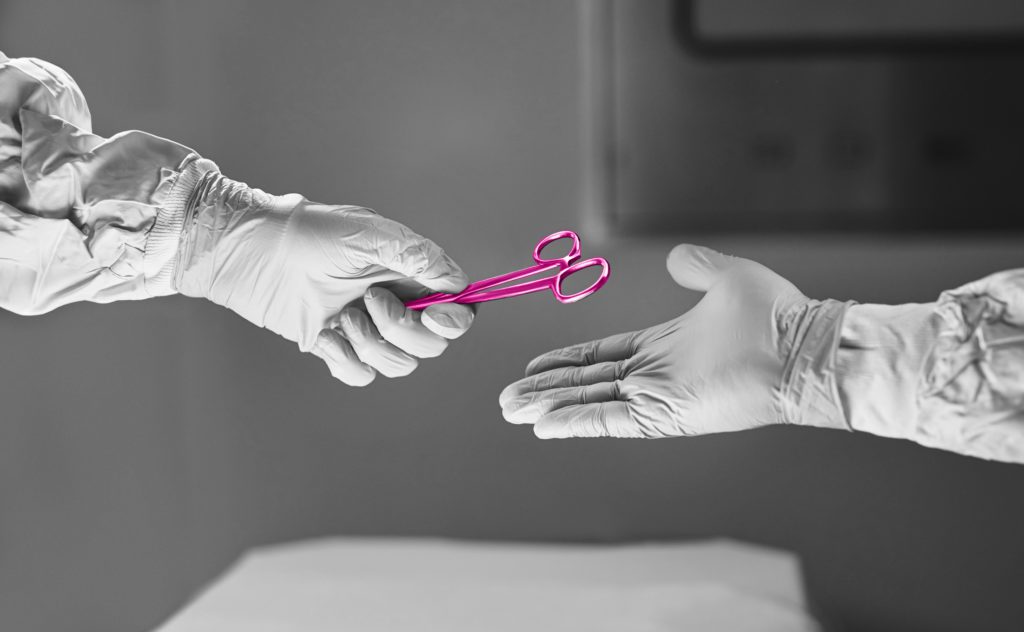 Image shows a doctors hand passing another doctor some scissors over an operating table. The scissors are painted pink to align with the brands colour scheme