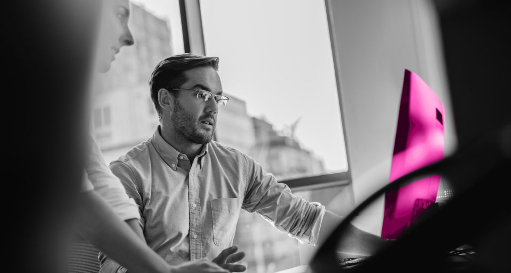 Image shows white man with dark hair and glasses at a computer desk with a woman looking over his shoulder at the computer.