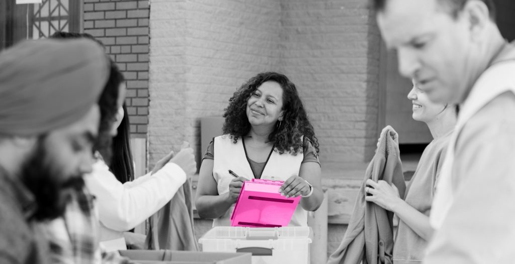 Image shows volunteer worker holding a clipboard and wearing a high visibility jacket interacting with other volunteers in an outside area. There are plastic boxes filled with donated clothes over which volunteers are conversing.