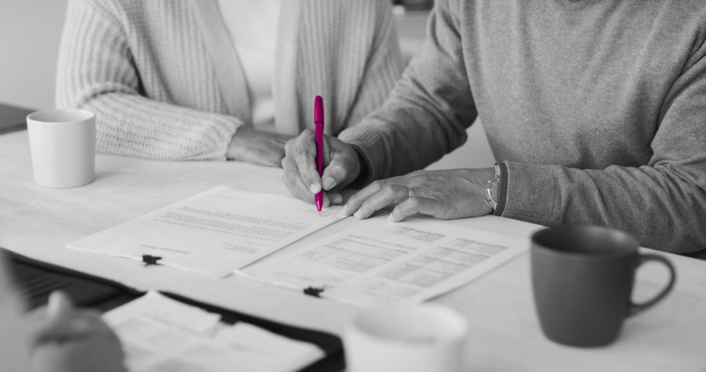 Image shows elderly couple sat at a table signing a document. There are mugs either side of the couple's arms and a lawyer sits opposite them.