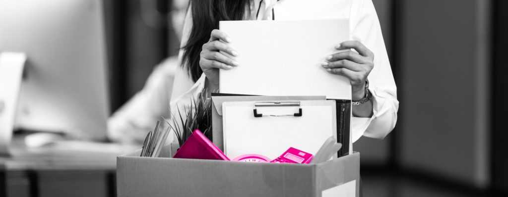 Image shows employee packing away files, an alarm clock and pencils into a cardboard box having been made redundant.